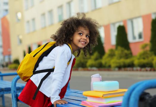 Young girl excitedly standing outside of an elementary school before the first day. She has her books next to her and is smiling by benches out front before starting her first day of a new grade. | Merry Maids®