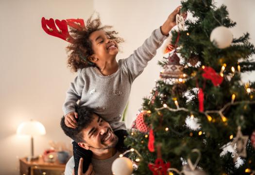 Beautiful young girl on her father's shoulders putting ornaments on the Christmas tree | Merry Maids