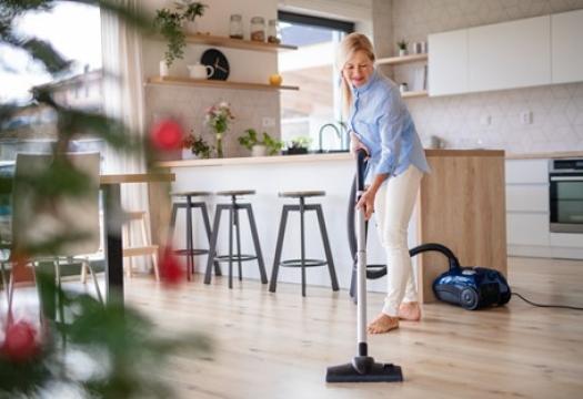 Woman smiling as she vacuums her hardwood floors in preparation of holiday company. She is tidying up and looking forward to her loved ones arriving. | Merry Maids®